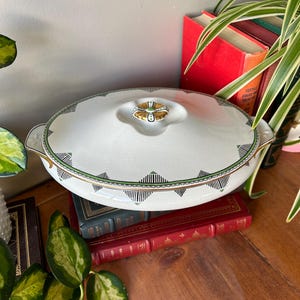 May include: Oval white serving dish with lid. The dish features a black and green geometric pattern around the rim and gold accents. The lid has a gold and green handle. The dish is sitting on a stack of books.