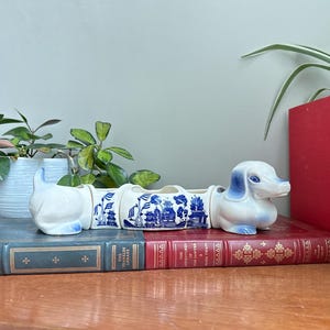 Peut inclure: Un cache-pot en céramique en forme de teckel composé de quatre segments reliés. La tête et la queue sont blanc uni avec des accents bleus, tandis que les deux sections centrales présentent un motif bleu et blanc de pagodes et d'arbres.