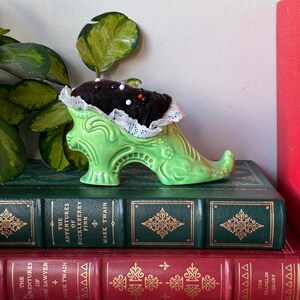 May include: A green ceramic shoe-shaped pin cushion with a dark brown cushion and white lace trim. The pin cushion is filled with colorful pins. It sits atop a stack of vintage books with gold lettering and detailing.