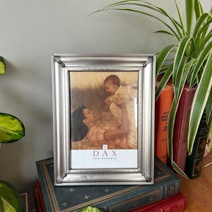 May include: Silver picture frame with a photo of a mother holding a baby. The frame has a decorative border and the text "DAX Photo Frames" is printed on the bottom.