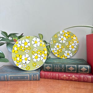 May include: Two round decorative coasters with a floral pattern. The coasters feature a light green background with white and yellow flowers. They are displayed on a wooden surface with vintage books and a potted plant.