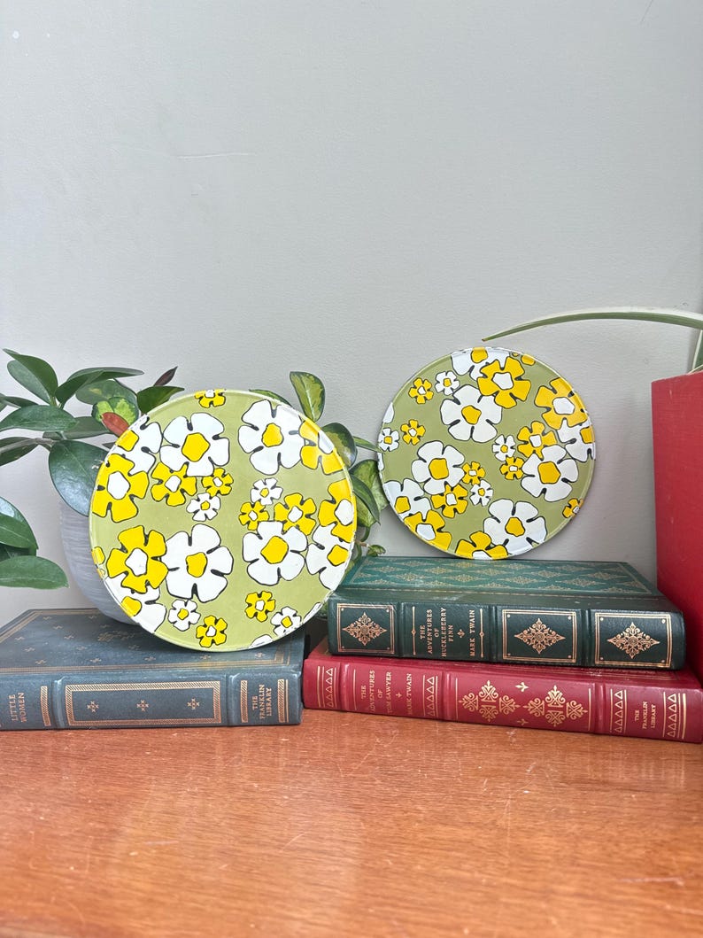 May include: Two round decorative coasters with a floral pattern. The coasters feature a light green background with white and yellow flowers. They are displayed on a wooden surface with vintage books and a potted plant.
