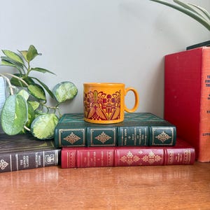 May include: An orange ceramic mug with a decorative design in red and yellow sits on a stack of vintage books. The books are bound in green, red, and brown leather, with gold lettering. A houseplant is visible in the background.