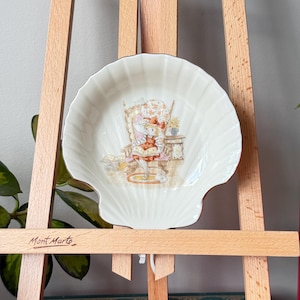 May include: A white, shell-shaped decorative dish with a detailed illustration of a girl reading in a chair. The dish has a gold trim and is displayed on a wooden easel. The easel has the text "Mont Marte" written on it.