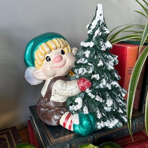 Könnte beinhalten: Eine Keramik-Weihnachtswichtelfigur, die einen grünen Weihnachtsbaum mit weißen Schneeakzenten hält. Der Wichtel trägt einen grünen Hut, eine braune Latzhose und weiß-rot gestreifte Socken.