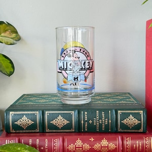 May include: A clear glass featuring a Mickey Mouse design with the text "MICKEY" in a pink and blue circle, with the years 1928 and 1988. The glass is set on top of two antique books with green and red covers.