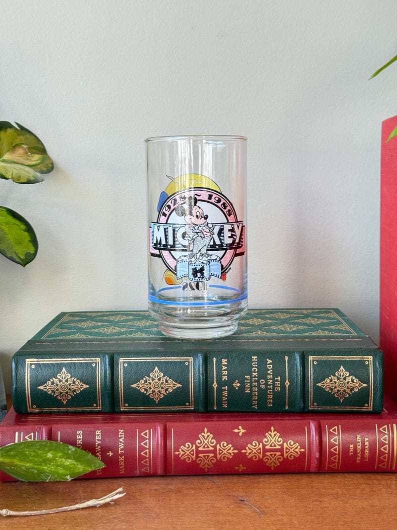 May include: A clear glass featuring a Mickey Mouse design with the text "MICKEY" in a pink and blue circle, with the years 1928 and 1988. The glass is set on top of two antique books with green and red covers.