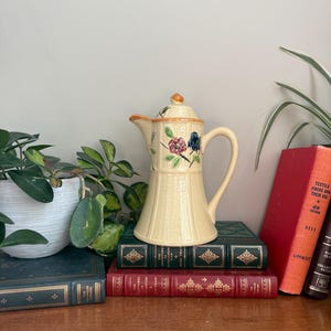 May include: Cream-colored vintage ceramic coffee pot with a lid, decorated with floral designs in pink, blue, and orange. The pot has a ribbed texture and a curved handle. A charming piece for kitchen decor.
