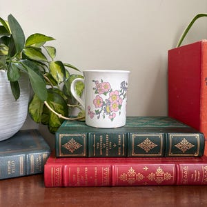 May include: A white ceramic mug featuring a floral pattern of pink blossoms with yellow centers and green leaves on a branch. The mug has a curved handle and is positioned on top of two stacked hardcover books with gold embossed lettering.