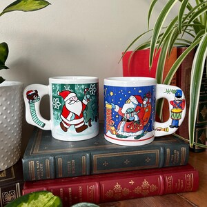 May include: Two white ceramic Christmas mugs with festive holiday designs. One mug features Santa Claus with a Christmas tree, the other features Santa Claus with a sack of gifts. Both mugs have a handle shaped like a Christmas character.