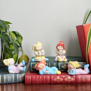 May include: Five small porcelain figurines of children with yarn-like hair in various poses. One sits with a puppy, one holds flowers, one lies on its stomach, and two are reclining. They wear pastel-coloured outfits including dresses and pyjamas.