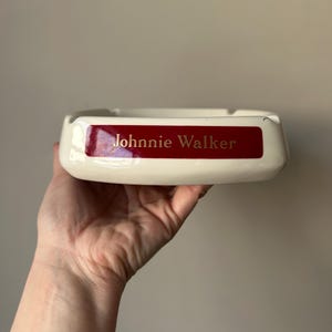 Op de afbeelding: Witte asbak van keramiek met een rood label waarop "Johnnie Walker" in gouden letters staat.