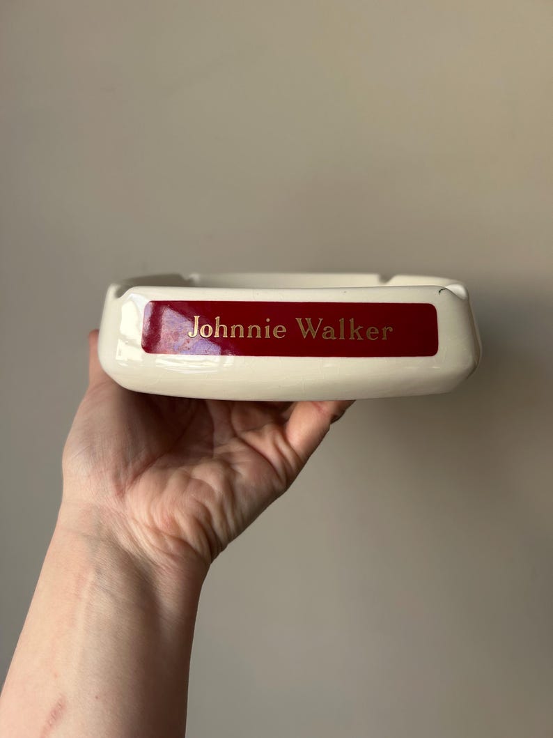 Op de afbeelding: Witte asbak van keramiek met een rood label waarop "Johnnie Walker" in gouden letters staat.