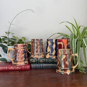 May include: Four ceramic beer steins with handles, each featuring a distinct hand-painted wavy pattern. One mug has red and blue swirls, another has brown and blue zigzags, one has bold blue stripes on tan, and the last has green and brown waves.
