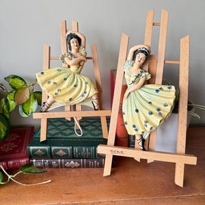 May include: Two decorative figurines of dancers in yellow dresses with blue floral accents, mounted on miniature wooden easels. The figurines have dark hair and are posed in a ballet-like stance. The easels are made of light-colored wood.