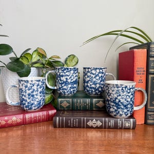 May include: Four ceramic mugs with a blue and white speckled design. The mugs are displayed on a wooden surface with antique books and potted plants. The mugs have a classic, farmhouse style.