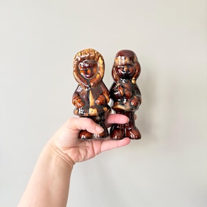 May include: Two ceramic figurines of people wearing brown and yellow winter coats and hats. The figurines are standing on a hand.