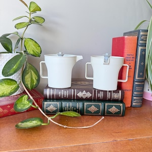 May include: Two white ceramic creamer pitchers with gray lids and handles. The pitchers are sitting on top of a stack of books.