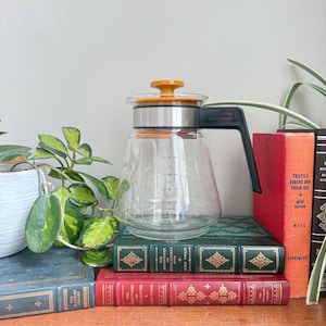 May include: A clear glass coffee maker with a black handle and an orange lid. The coffee maker is sitting on top of a stack of vintage books with gold lettering. Green plants are in the background.