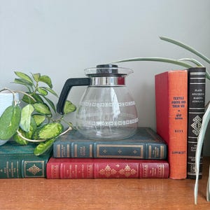 May include: A clear glass coffee carafe with a black handle and lid, decorated with a white geometric pattern. The carafe sits atop a stack of vintage books in red, green, and blue hues, with a plant in the background.