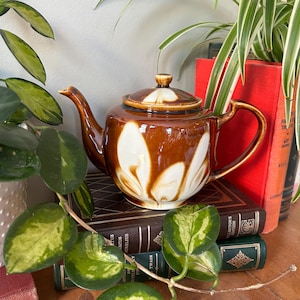 May include: A brown and white ceramic teapot with a floral design sits on top of a stack of books. The teapot has a lid and a spout. The books are covered in brown leather and have gold lettering on the spines.