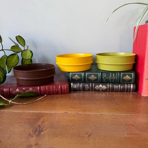 Puede incluir: Tres cuencos redondos y coloridos, en marrón, amarillo y verde, apilados sobre una colección de libros antiguos. Los cuencos tienen un diseño sencillo y moderno con un borde ligeramente elevado. Los libros son de varios colores y tamaños.