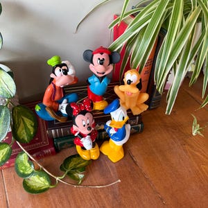 Könnte beinhalten: Eine Sammlung farbenfroher Disney-Charakterfiguren, darunter Micky Maus, Minnie Maus, Donald Duck, Goofy und Pluto. Die Figuren stehen auf einer Holzoberfläche, mit Büchern und Grünzeug im Hintergrund.