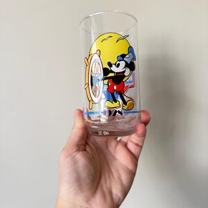 May include: Clear glass tumbler featuring a colorful graphic of Mickey Mouse at the helm of a ship, with a yellow sun in the background. The design includes the year "1928" at the bottom. The glass is held in a hand.