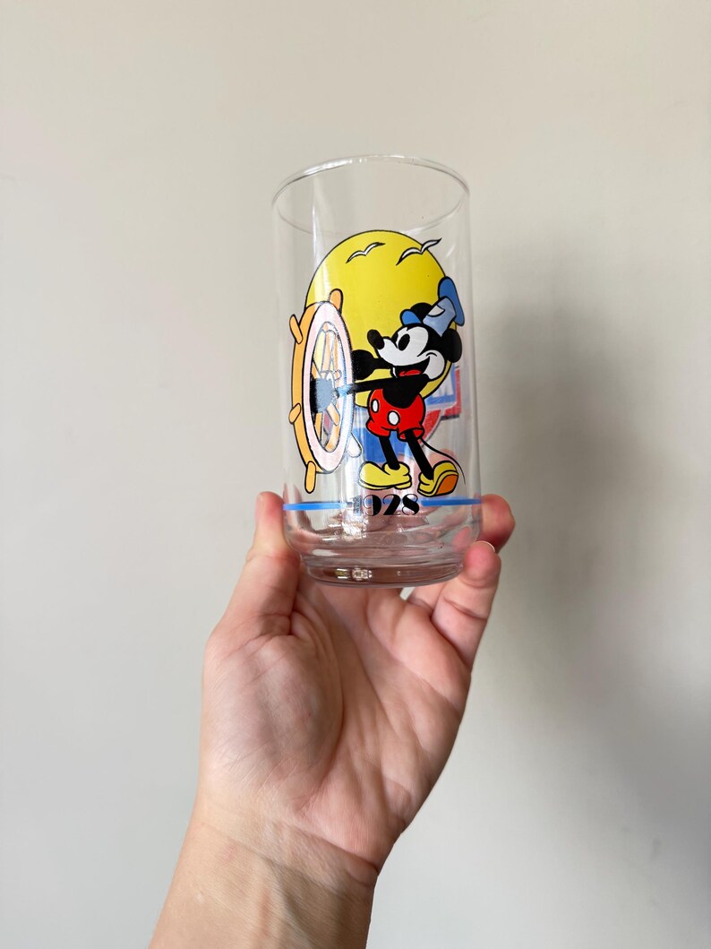 May include: Clear glass tumbler featuring a colorful graphic of Mickey Mouse at the helm of a ship, with a yellow sun in the background. The design includes the year "1928" at the bottom. The glass is held in a hand.