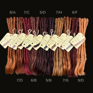 Könnte beinhalten: Eine Auswahl an Dreadlocks in verschiedenen Braun-, Rot- und Auburn-Tönen, in einer Reihe angeordnet. Jedes Bündel ist mit einem Farbcode und einem Etikett mit der Aufschrift "DREAM HUMAN HAIR" versehen. Der Hintergrund ist schwarz.