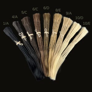 May include: A collection of dreadlock extensions in various shades, from dark black to light blonde. Each bundle is tied with twine and labeled with a color code. The background is a solid black.