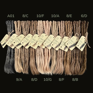 Könnte beinhalten: Eine Sammlung von Dreadlock-Extensions in verschiedenen Braun-, Blond- und Schwarztönen. Jedes Bündel ist mit einem Etikett mit der Aufschrift "DREAM HUMAN HAIR" versehen. Die Dreadlocks sind vor einem schwarzen Hintergrund angeordnet, um die verschiedenen Farboptionen zu präsentieren.