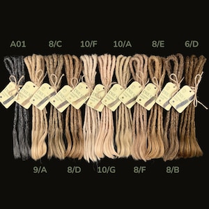 May include: A collection of dreadlocks in various shades of brown, blonde, and black, each bundled and labeled with tags that read "Dreads Human Hair". The dreadlocks are arranged against a black background, showcasing a range of colors and styles.