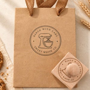 May include: A brown paper gift bag with brown ribbon handles, featuring a circular stamp design of a stand mixer and the text Baked With Care Sweet Whisk Co. A square wooden rubber stamp with the same logo sits next to the bag.