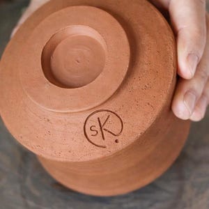 May include: A terracotta pottery item, possibly a small bowl or cup, held by a hand. The base has a circular design and a stamped "sK" logo. The unglazed clay has a warm, earthy tone, typical of earthenware.