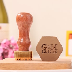 May include: A wooden handled stamp with a brass base and a hexagonal stamp with the initials "G&M" and the date "13.12.25" engraved on it. The stamp is on a wooden surface.