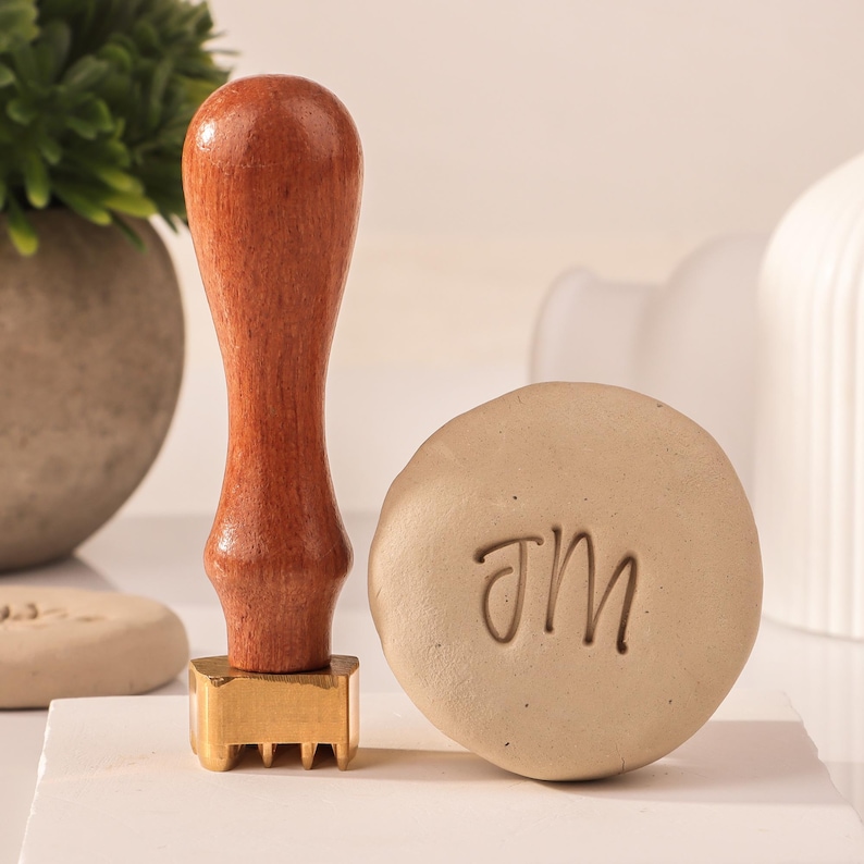 May include: A wooden handle stamp with a brass head and a decorative design. The stamp is used to create an imprint of the letters "JM" on a beige surface.