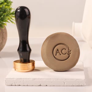 May include: A wooden stamp with a black handle and a brass base. The stamp features a circular design with the initials "AC" and a wheat stalk. The stamp is on a white surface.
