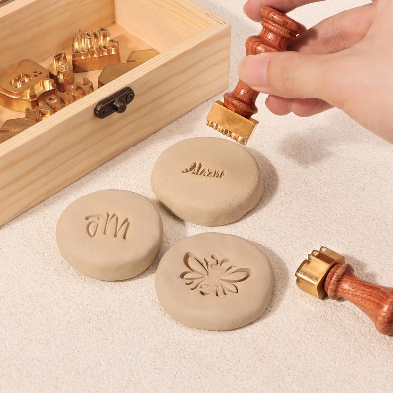May include: A set of three clay circles with different designs stamped into them using brass stamps. The stamps are shown in a wooden box with a variety of other stamps. The designs include the words "AM" and "Alexa" and a bee.