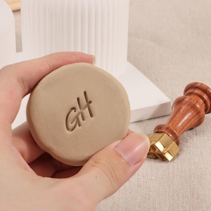 May include: A close-up of a brown clay disc with the initials "GH" stamped into the surface. A wooden and brass stamp with the same initials is visible in the background.