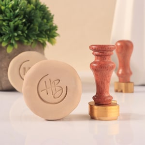 Op de afbeelding: Een verzameling waszegels en stempels. De zegels zijn rond en beige met een reliëfontwerp. De stempels hebben houten handvatten en messing bases. Eén zegel heeft de initialen "HB" in reliëf.
