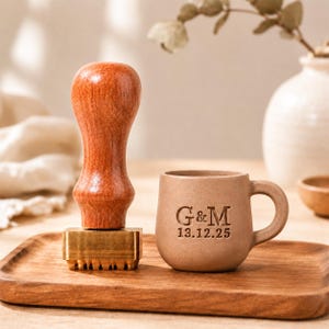Puede incluir: Un sello de cera con mango de madera de color marrón rojizo liso y cabeza de latón rectangular, junto a una pequeña taza de cerámica de color beige mate con el texto grabado G&M y la fecha 13.12.25 en su superficie frontal.