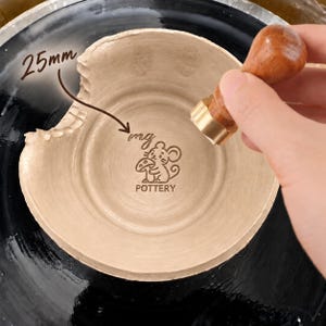 Może przedstawiać: Jasnobeżowa miska ceramiczna z teksturowanym, falistym brzegiem i wytłoczonym wzorem na środku przedstawiającym mysz trzymającą grzyba oraz napis mg POTTERY. Drewniana pieczątka z mosiężną podstawą odciska się w glinie, z oznaczeniem 25mm.