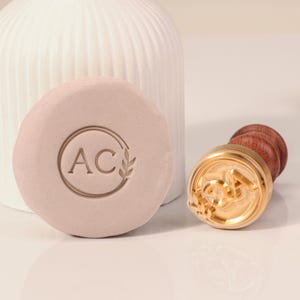 Puede incluir: Un sello de metal dorado con mango de madera. El sello presenta las letras "AC" en un círculo con un diseño de hoja. El sello se utiliza para crear una impresión en una superficie rosa claro.