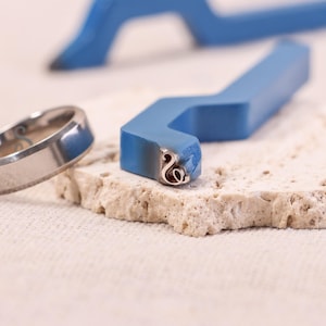 Può includere: Primo piano di strumenti per la creazione di gioielli, tra cui un anello d'argento e un timbro metallico blu con un design decorativo. Il timbro è appoggiato su una superficie strutturata bianco sporco. L'anello ha una finitura lucida.