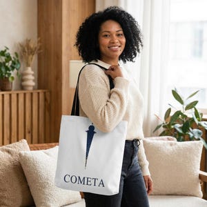May include: White tote bag with black straps featuring a navy blue design and the word "COMETA" in navy blue. The design is a stylized illustration. The bag is held by a person.