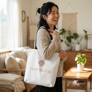 Könnte beinhalten: Weiße Tragetasche mit einem rosa Muschelmotiv und dem Wort "CONCHIGLIA". Die Tasche wird von einer Person mit einem beigen Pullover getragen. Die Tasche hat schwarze Träger.