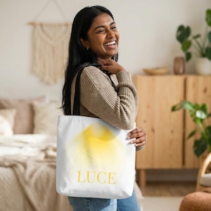 Könnte beinhalten: Weiße Tragetasche mit gelbem, abstraktem Design und dem Wort "LUCE" in goldfarbenen Buchstaben. Die Tasche hat schwarze Träger.