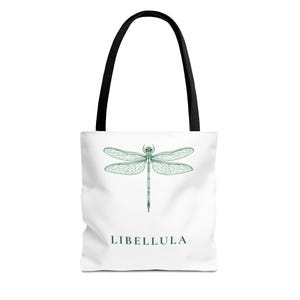 Könnte beinhalten: Weiße Tragetasche mit schwarzen Griffen, mit einer grünen Libellenillustration. Das Wort "LIBELLULA" ist grün darunter gedruckt. Die Tasche ist zum Tragen von Gegenständen konzipiert.