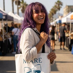 Könnte beinhalten: Eine Frau mit lila Haaren lächelt und hält eine weiße Tragetasche mit dem Wort "MARE" und einem blauen Schleifendesign. Die Tasche hat schwarze Träger. Der Hintergrund zeigt einen sonnigen Markt im Freien mit Verkaufsständen und Menschen.